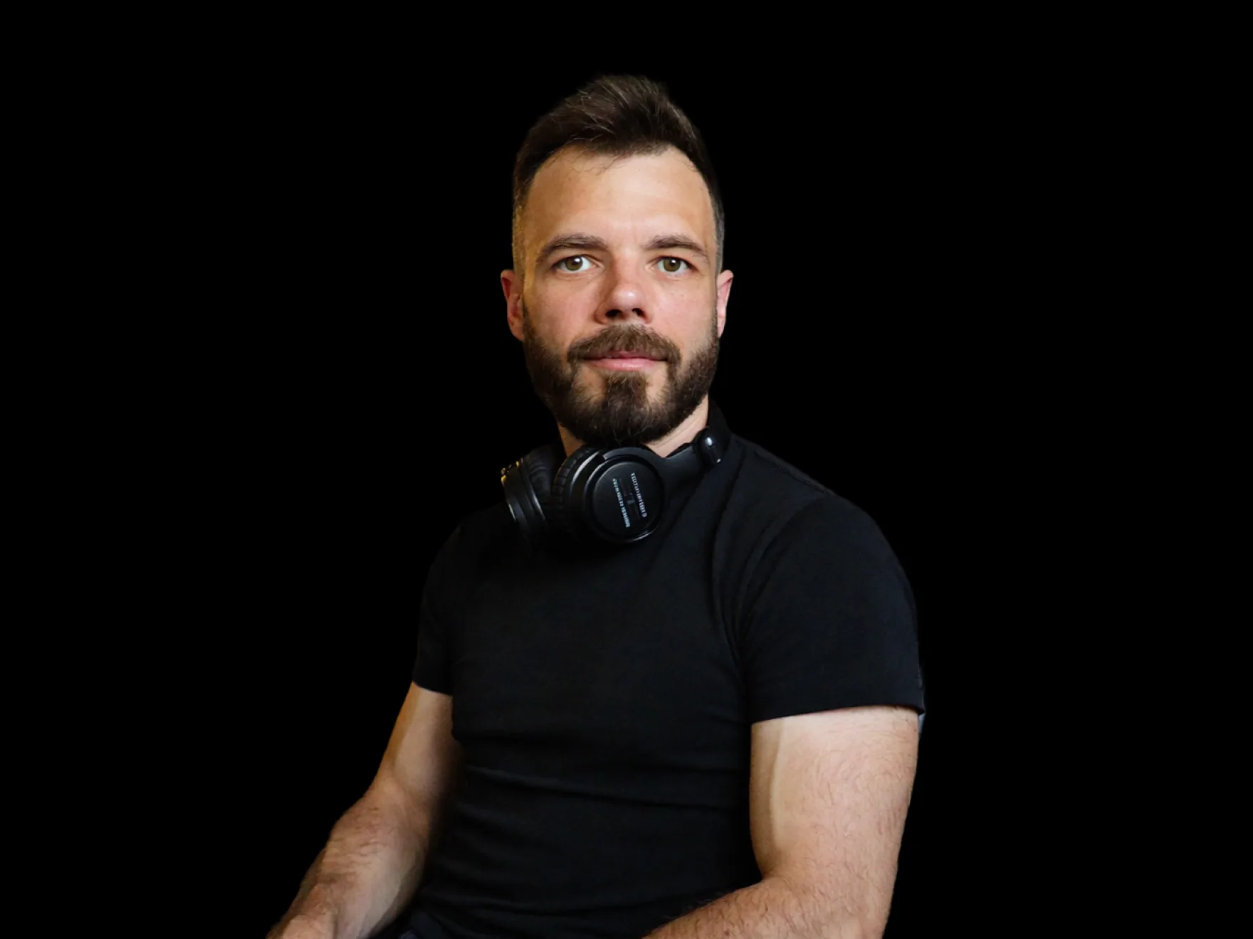 Russ Hepton in a dark studio, plain black t-shirt, headphones around his neck, looking down the lens