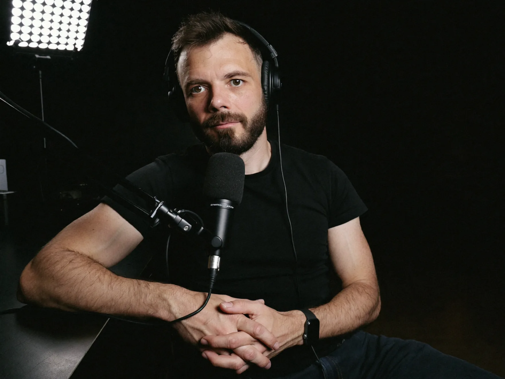 Russ Hepton seated at a desk in a darkened studio, headphones on and hands clasped behind a microphone