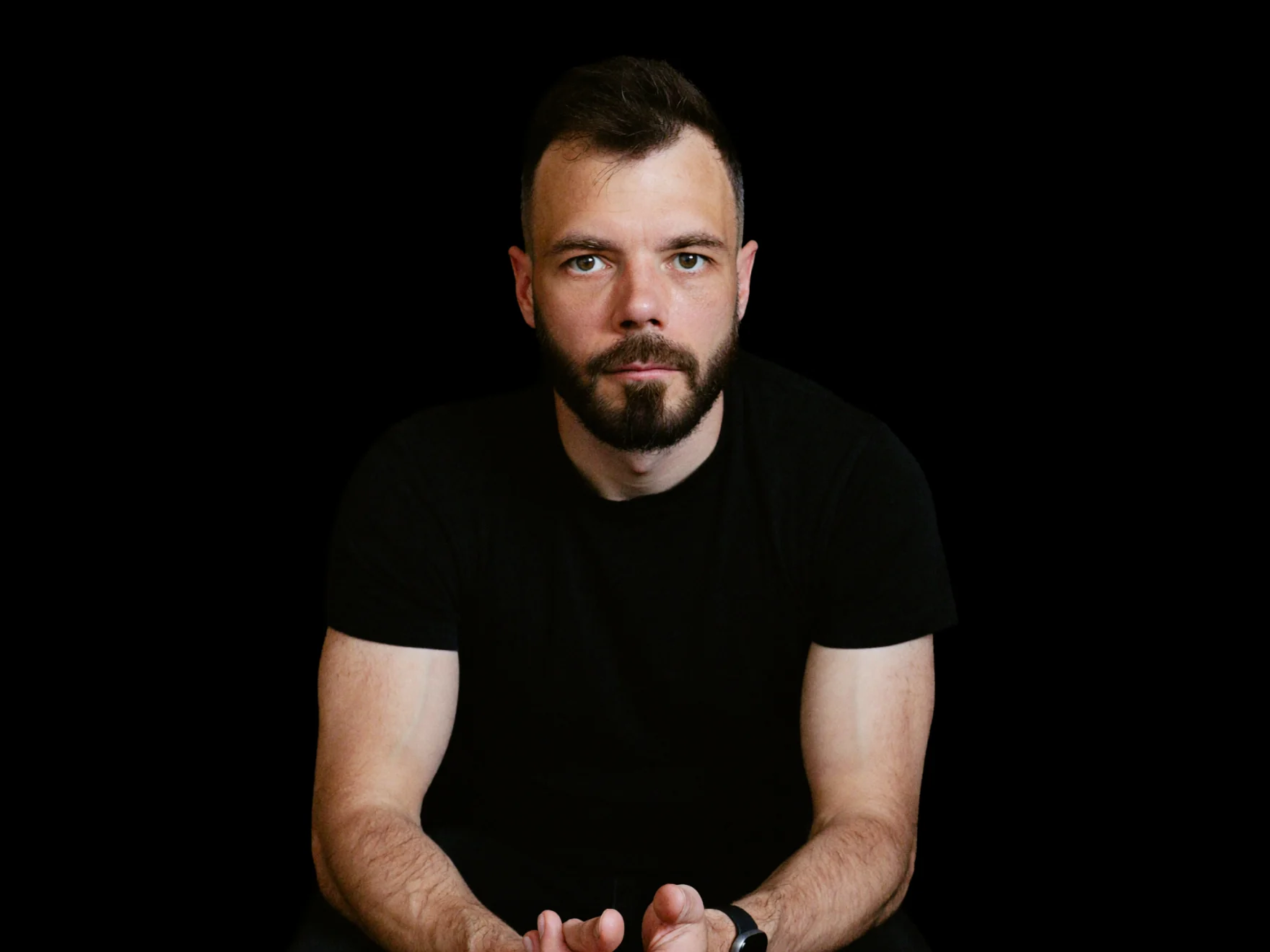 Russ Hepton seated on a stool in a dark studio, plain black t-shirt, looking straight down the lens